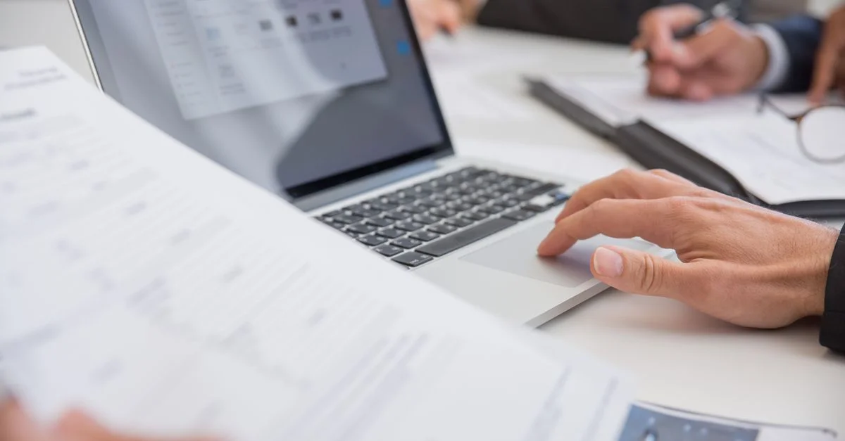 Gros plan sur des mains travaillant sur des documents et un ordinateur portable dans un bureau, illustrant le travail d'équipe et la productivité.