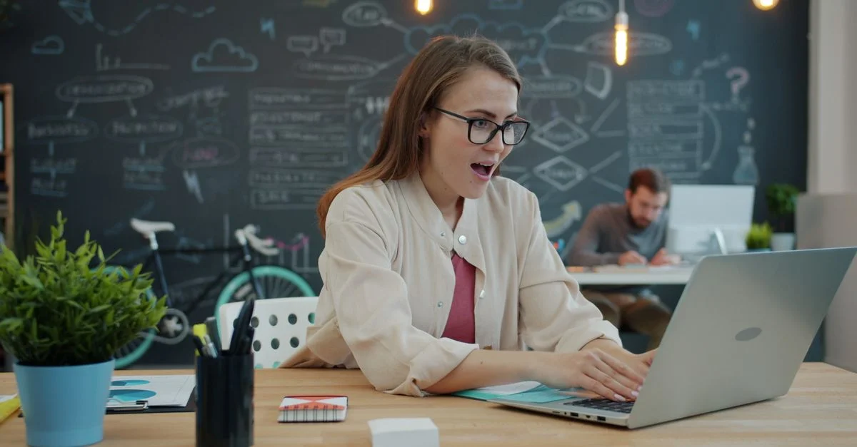 Femme souriante au bureau, fascinée par son ordinateur portable, espace de travail créatif avec tableau noir en arrière-plan.