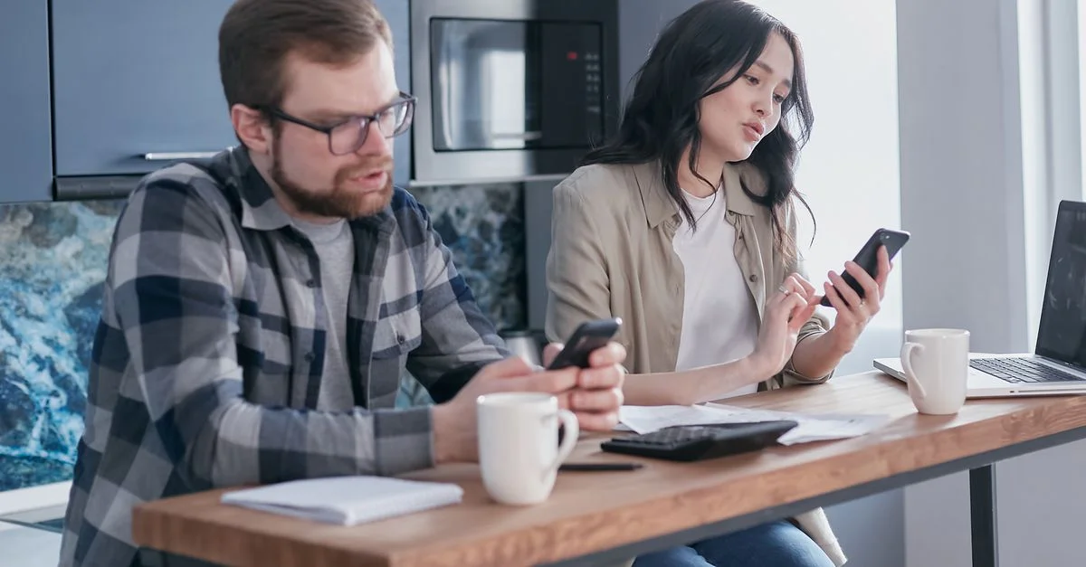 Jeune couple gérant leurs finances avec des smartphones et un ordinateur portable dans une cuisine moderne.