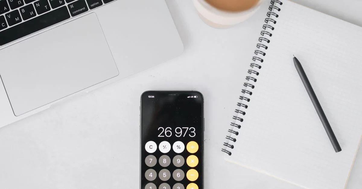 Vue de dessus d'un ordinateur portable, d'un smartphone affichant une calculatrice, d'un carnet, d'un stylo et d'un café sur une table blanche.