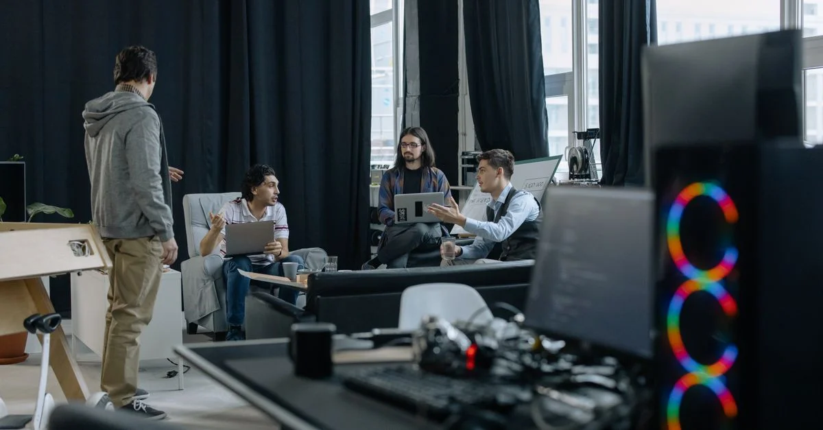 Un groupe de professionnels en réunion informelle dans un bureau moderne, discutant de projets sur des ordinateurs portables et des tablettes.