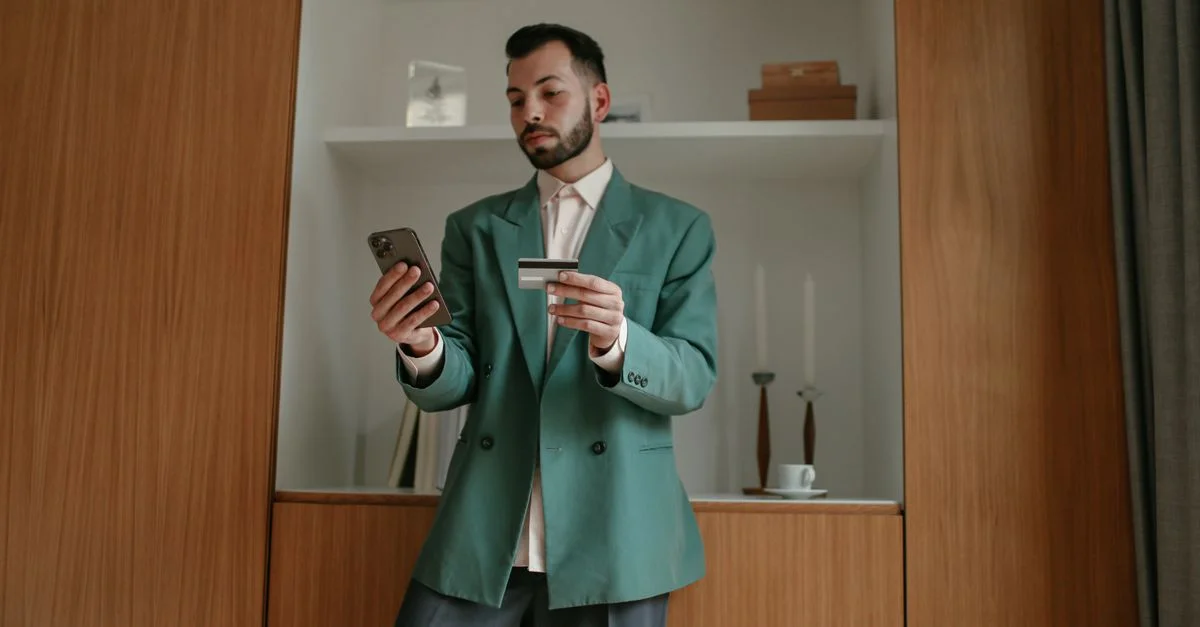 Un homme barbu en costume vert utilise un smartphone et une carte de crédit à l'intérieur, mettant en avant la technologie moderne et le style de vie.